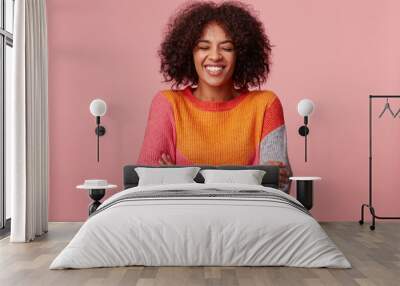 Happy joyful attractive African American with an afro hairstyle closed eyes and laughs from something funny, stand with arms crossed, wearing colorful sweater, isolated on pink Wall mural