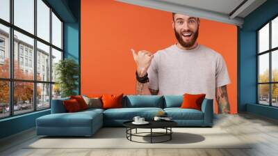 Happy pretty bearded tattooed man with trendy haircut looking at camera with wide smile and rounding his blue eyes, pointing aside with raised thumb while posing over orange background Wall mural