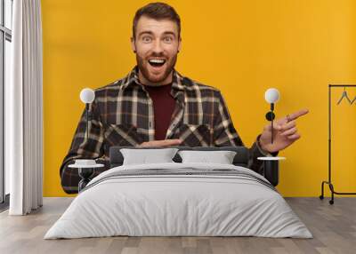 Happy wondered young man in plaid shirt with beard and opened mouth looks amazed and pointing away at empty space by two fingers over yellow background Wall mural