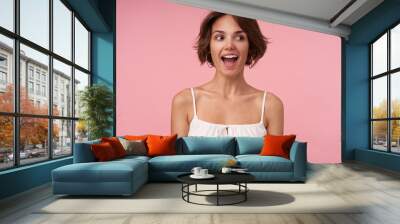 Joyful attractive brunette female with short haircut looking aside with wide happy smile, keeping hands along her body, posing over pink background, being in high spirit Wall mural