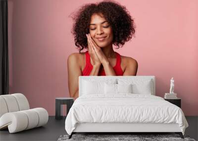 Pleased charming African American woman with afro hairstyle feels pleasure, touches her face with palm, closed eyes, rejoices, folded hands as sleepy, wearing red singlet, isolated on pink background Wall mural