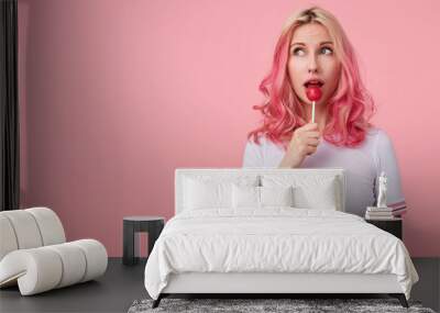 Portrait of attractive young tattooed lady with pink curly hair looking aside dreamily, posing over pink background in casual white t-shirt and licking candy on stick Wall mural