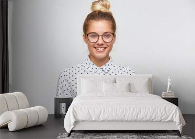 Portrait of cheerful beautiful young woman with bun wears polka dot shirt and spectacles feels confident and looks to the camera isolated over white background Wall mural