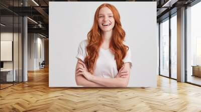 Portrait of cheerful pretty redhead young woman with long wavy hair and freckles wears t shirt keeps hands folded and laughing isolated over white background Wall mural