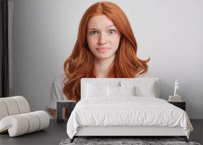 Portrait of confused astonished young woman with long wavy red hair and freckles wears t shirt feels embarrassed and looks directly in camera isolated over white background Wall mural