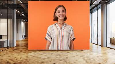 Portrait of girl with brown short haircut and braces for teeth, pierced nose, wearing striped shirt. Young smiling girl watching camera against orange background Wall mural