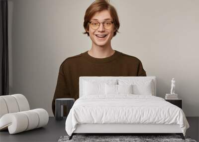 Portrait of handsome, cheerful male with blond hair. Wearing brown sweater, glasses and has braces. Holding a smartphone and smiling. Watching at the camera isolated over grey background Wall mural