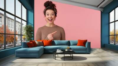 Portrait of happy amazed young beautiful lady with curly dark hair, heard the cool news, broadly smiling, looking at the camera, pointing with finger to copy space isolated over pink background. Wall mural