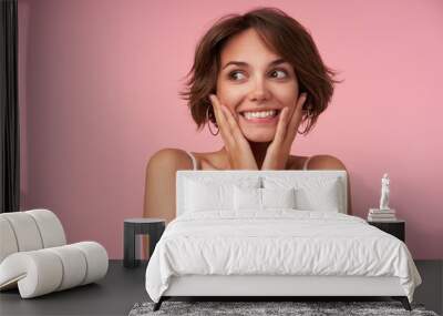 Portrait of happy attractive young woman with short brown hair holding palms on her cheeks and looking aside with charming smile, wearing white top over pink background Wall mural