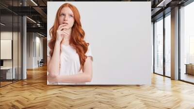 Portrait of pensive attractive redhead young woman with long wavy hair and freckles wears t shirt touching her chin and thinking about future isolated over white background Wall mural