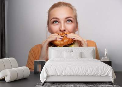 Portrait of pleased young lovely blonde woman with casual hairstyle eating fresh hamburger with great appetite and looking cheerfully upwards, posing over white background Wall mural