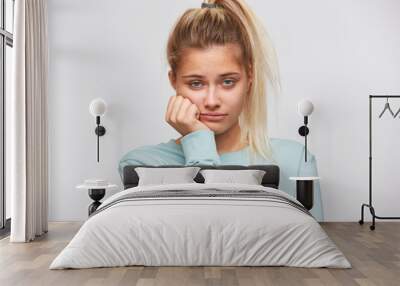 Portrait of unhappy disappointed young woman with blonde hair and ponytail wears blue sweatshirt looks bored and sad isolated over white background Wall mural