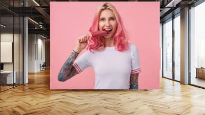 Portrait of young beautiful pink-haired girl with tattooed hands, wearing a white t-shirt, looking at the camera and trying to bite off the ice cream, standing over pink background. Wall mural