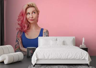 Portrait of young thinking cute lady with pink hair, wears blue shirt, stands over pink background copy space with crossed arms, looks up thoughtfully. People and emoyion concept. Wall mural