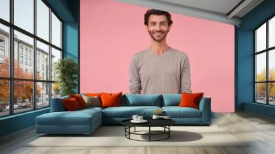 Studio shot of beautiful bearded male with dark hair standing over pink background, wearing casual clothes, looking at camera cheerfully with hands down Wall mural