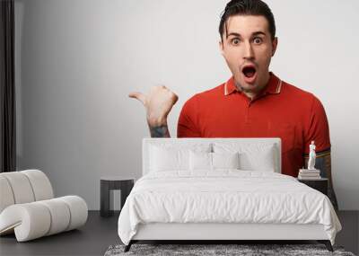 Stylish man fascinated and excited. Close up of amazed attractive young male in red polo pointing aside with dropped jaw, standing over white wall Wall mural