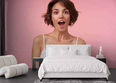 Surprised pretty young brunette woman with short haircut looking at camera with wide eyes and mouth opened, posing over pink background with hands down Wall mural