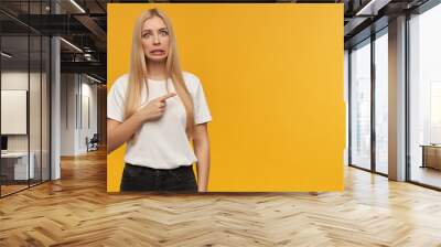 Teenage girl, unhappy looking woman with blond long hair. Wearing white t-shirt and black jeans. People and emotion concept. pointing to the right at copy space with a disgust facial expression Wall mural