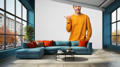 Young attractive handsome guy looks delighted gladden amazed, mouth open, smiling, like dream came true, big discount in shop,wears orange sweatshirt, points with thumb finger on a blank copyspace Wall mural