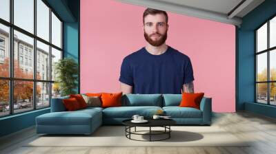 Young attractive red-bearded guy with blue eyes, wearing a blue t-shirt, looking at the camera with a calm facial expression isolated over pink background. Wall mural