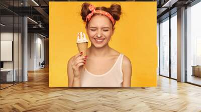 Young lady, pretty ginger woman with two buns. Wearing white tank top and red doted hairband. Holding ice cream and have it on her nose, smiling. Stand isolated over yellow background Wall mural