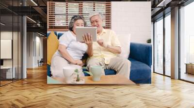 Asian elderly couple using tablet watching TV in living room at home, couple enjoy love moment while lying on sofa when relaxed at home. Enjoying time lifestyle senior family at home concept. Wall mural