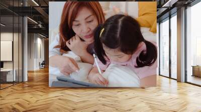 Asian grandmother using tablet read fairy tales to granddaughter at home. Senior Chinese, grandma happy relax with young girl before bedtime lying on bed in bedroom at home at night concept. Wall mural