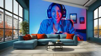 Happy asia girl gamer wear headphone set and joystick controller talk with friend feel fun and excited with competition online game in neon living room home studio at night, Home quarantine activity. Wall mural