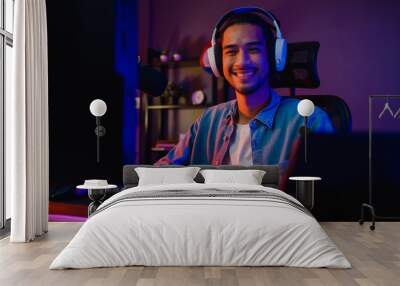 Happy asia guy gamer wear headphone participation play video game colorful neon lights computer and smiling look at camera in living room at night modern house. Esport streaming game online. Wall mural