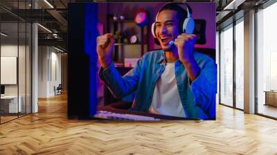 happy asia guy gamer wear headphone participation play video game colorful neon lights computer in l Wall mural