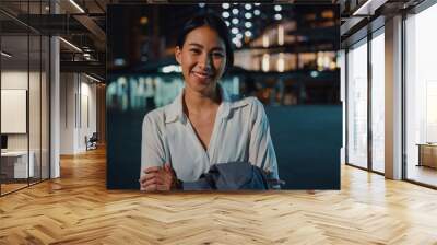 Successful young Asia businesswoman in fashion office clothes smiling and looking at camera while happy standing alone outdoors in urban modern city at night. Business on the go and commuter concept. Wall mural