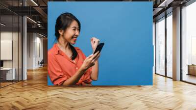 Surprised young Asia lady using mobile phone with positive expression, smiles broadly, dressed in casual clothing and stand isolated on blue background. Happy adorable glad woman rejoices success. Wall mural