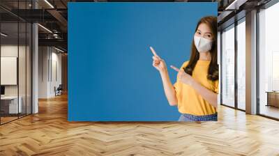 Young Asia girl wear medical face mask shows something at blank space with dressed in casual cloth and look at camera. Social distancing, quarantine for corona virus. Panoramic banner blue background. Wall mural