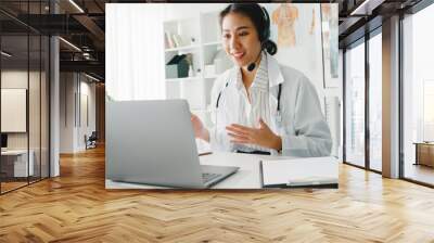 Young Asia lady doctor in white medical uniform with stethoscope using computer laptop talking video conference call with patient at desk in health clinic or hospital. Consulting and therapy concept. Wall mural