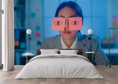 Young Asian businesswoman sitting on desk overworked tired sleep over a laptop at office at night. Exhausted burnout lady with two post-it over her eyes, adhesive notes on face sleeping at workplace. Wall mural