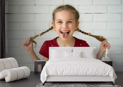 Having hair in two plaits. Adorable funny girl holding pigtails hair on a brick wall. Little cute girl with long blonde hair outdoor fooling around, showing tongue Wall mural