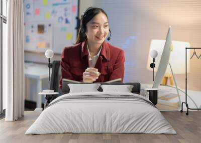 Asian businesswoman happily working late in her well-lit office, smiling during a video call, surrounded by technology and paperwork, showcasing her dedication to her career Wall mural