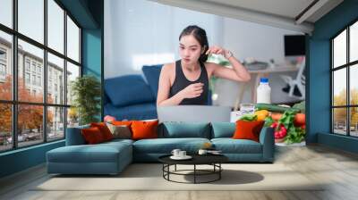 Asian nutritionist wearing sportswear is gesturing while giving online diet consultation from her home office with healthy food, milk, and water on the table Wall mural