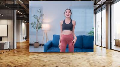 Asian woman in sportswear exercises happily at home, surrounded by furniture and plants, basking in natural light. Her smile shows dedication to wellness Wall mural