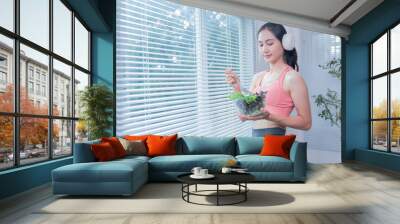 Happy asian fitness sportswoman eating healthy salad and listening to music with headphone in living room at home, healthy food and healthcare concept Wall mural