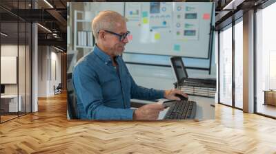 Smiling mature businessman holding credit card and using computer for online shopping at workplace, senior man purchasing goods or services on internet, using e-commerce platform Wall mural