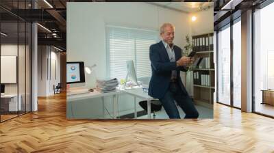 Smiling senior manager using smartphone in modern office with computer showing charts and graphs on screen, indicating successful business operations Wall mural