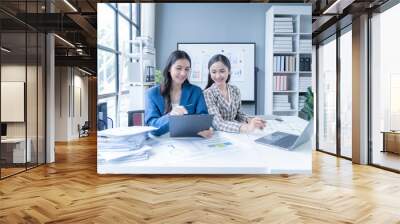 Two asian businesswomen are having a meeting and working together on a new project at their office desk using a laptop computer and analyzing documents Wall mural