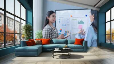 Two businesswomen are having a discussion about business strategy based on data displayed on charts and graphs on a whiteboard Wall mural