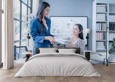 Two businesswomen in a modern office collaborate on documents, analyzing data and discussing strategies in a bright, positive atmosphere filled with teamwork Wall mural