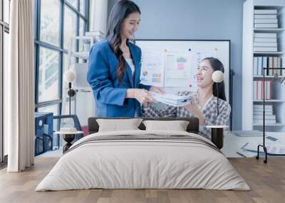 Two smiling businesswomen exchange documents in a modern office, showcasing successful teamwork in a professional setting. Bright, positive atmosphere with laptops and large windows Wall mural