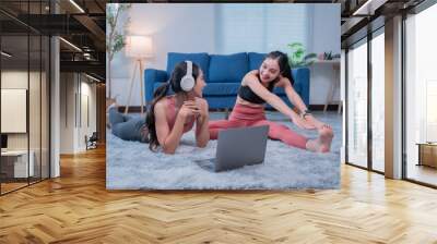 Two sporty girls wearing headphones and sportswear are stretching muscles watching online fitness tutorials on a laptop at home Wall mural