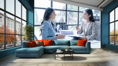 Two young businesswomen in a modern office, analyzing financial charts and reports, collaborating on strategies, and planning together in the bright, natural light Wall mural