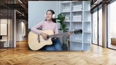 weekend day asian girl is sitting on couch blowing off steam by singing playing guitar at home. woman holding microphone with exaggerated body movement is having happy and crazy time alone. Wall mural