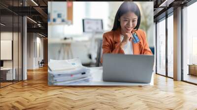 Young asian businesswoman in a modern office, multitasking with a laptop and credit card while shopping online Wall mural